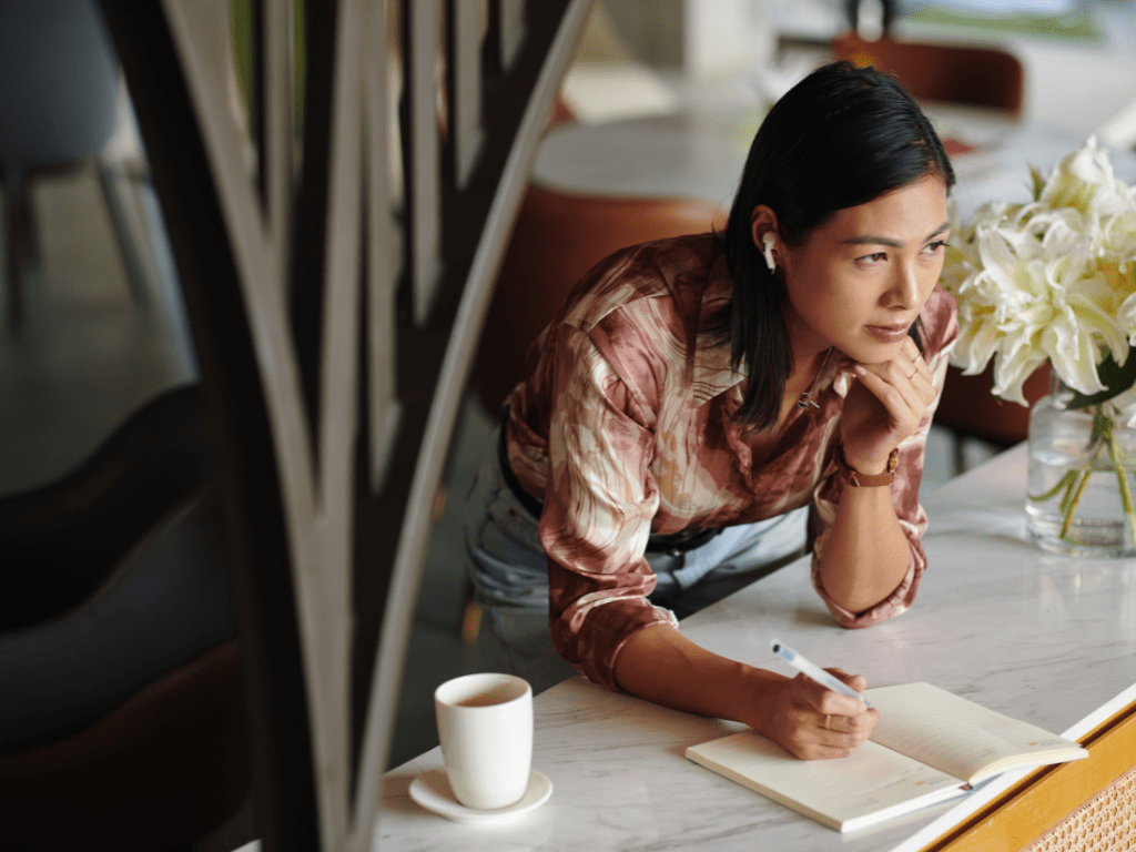Frau lehnt an einem Tisch, schreibt in ein Notizbuch und denkt nach