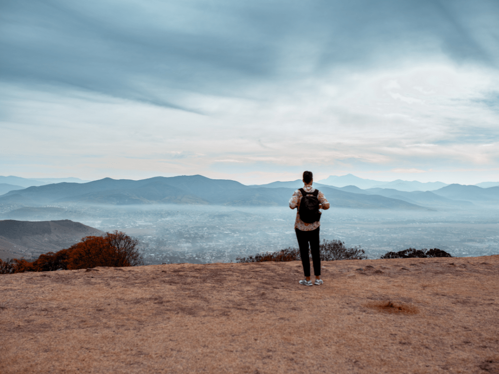 Person steht auf einem Felsen und blickt über eine weite Berglandschaft – Moment des Innehaltens beim Imposter-Syndrom.
